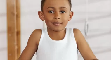 A young boy stands between parallel bars in a gymnastics studio, smiling.