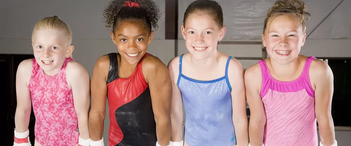 Four young gymnasts hold themselves up on the bar, wearing leotards and smiling for the photo. There are three Caucasian girls, and one African American girl. They are all happy to be participating in the girls gymnastics youth program at the YMCA.