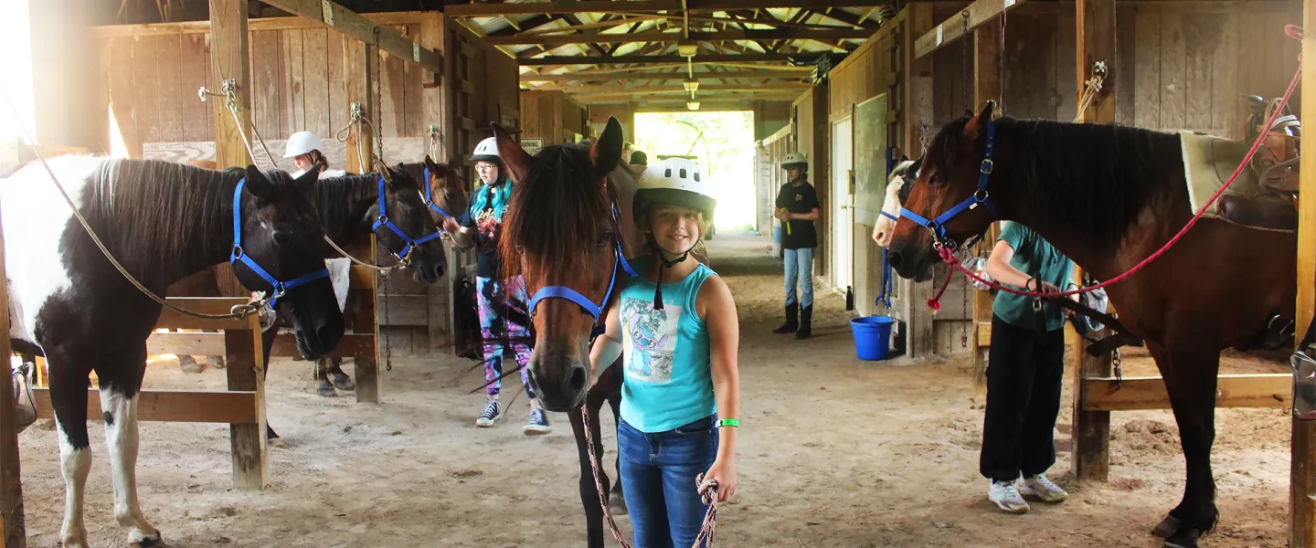 A girl participating in a YMCA Camp Lakewood Equestrian Specialty Camp smiles at the Triangle Ranch Barn with her camp horse Rocco
