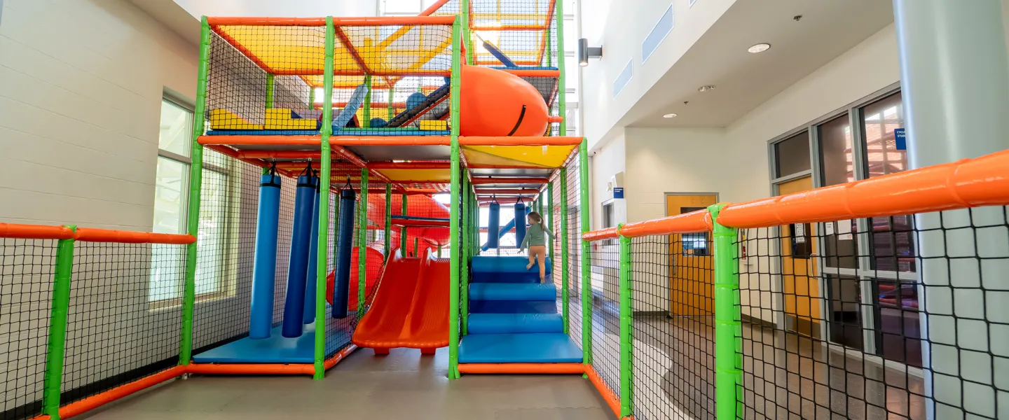 A massive, multi-level indoor play structure for children featuring orange and green padding, safety netting, blue hanging bolsters, and a bright orange tunnel slide.