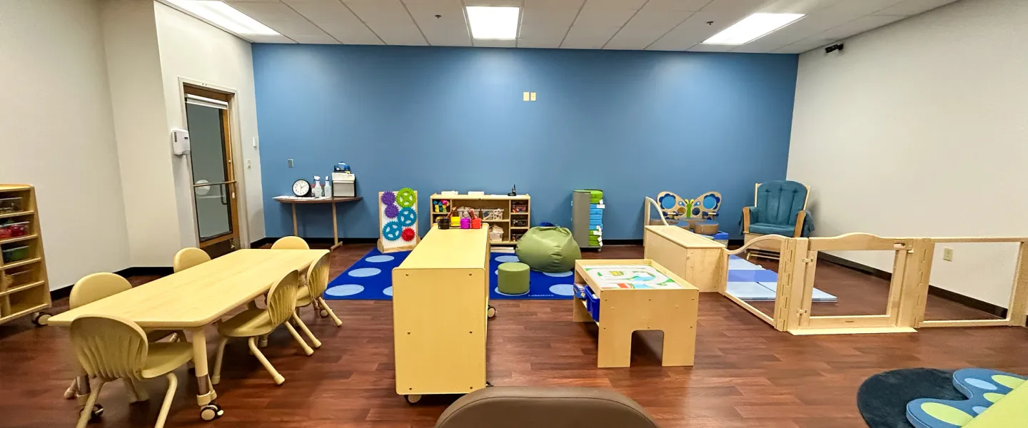 The center of the Child Watch room showing a blue feature wall with gear-themed wall toys, a wooden activity table, and a brown child-sized armchair.