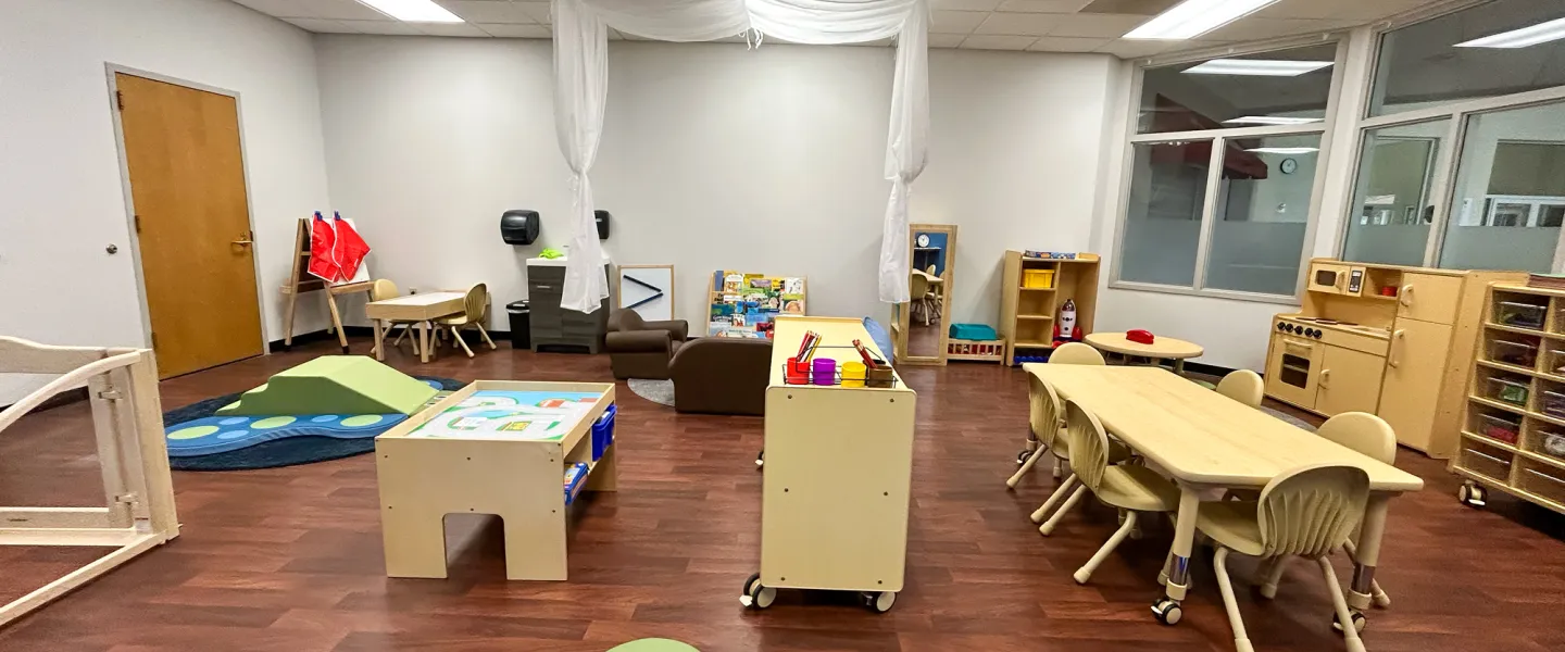View of the Child Watch play area featuring a wooden play kitchen, a large reading table with chairs, and a whimsical white fabric canopy draped from the ceiling.