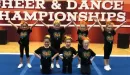 Thumbnail: An image of young, Caucasian girls competing in a cheerleading competition at the YMCA. The girls are wearing orange bows on their heads, and matching black tops paired with black pants.